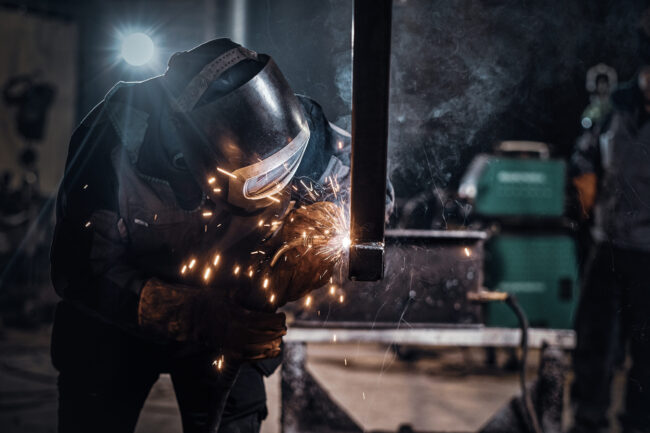 Schweißer mit Schutzhelm bearbeitet ein Metallbauteil in der Fertigung von Sondermaschinen, Funkenflug während des Schweißvorgangs.