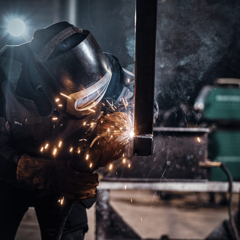 Schweißer mit Schutzhelm bearbeitet ein Metallbauteil in der Fertigung von Sondermaschinen, Funkenflug während des Schweißvorgangs.
