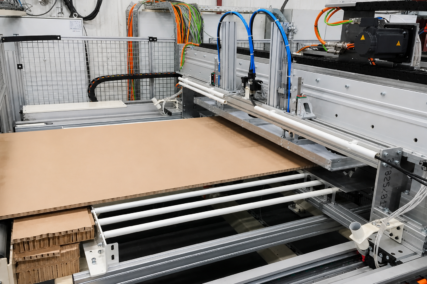 Detailaufnahme einer automatisierten Anlage zur Wabenplattenproduktion mit Zuführung einer Kartonplatte über Rollenbahn sowie mechanischen und pneumatischen Komponenten in einer industriellen Umgebung