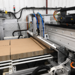 Detailaufnahme einer automatisierten Anlage zur Wabenplattenproduktion mit Bearbeitungseinheit und Zuführung von Kartonplatten über eine Rollenbahn, inklusive mechanischer und pneumatischer Komponenten in industrieller Umgebung