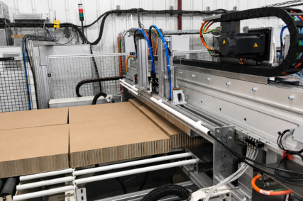 Detailaufnahme einer automatisierten Anlage zur Wabenplattenproduktion mit Bearbeitungseinheit und Zuführung von Kartonplatten über eine Rollenbahn, inklusive mechanischer und pneumatischer Komponenten in industrieller Umgebung