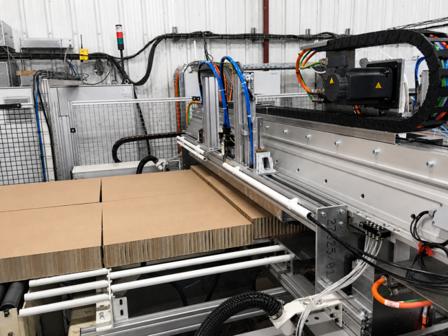 Detailaufnahme einer automatisierten Anlage zur Wabenplattenproduktion mit Bearbeitungseinheit und Zuführung von Kartonplatten über eine Rollenbahn, inklusive mechanischer und pneumatischer Komponenten in industrieller Umgebung
